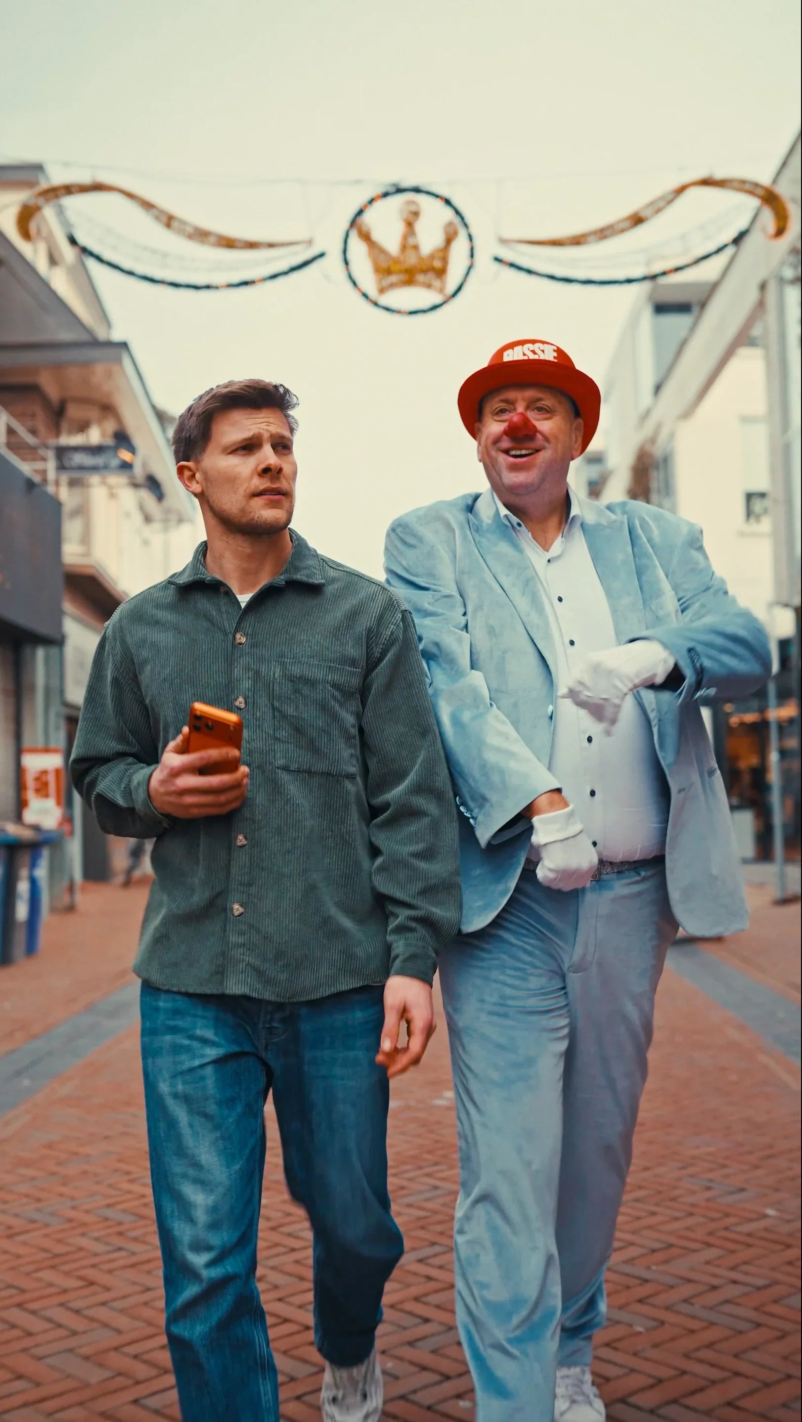 Twee mannen lopen buiten; één houdt een oranje iPhone, de ander draagt clownsmuts en rode neus