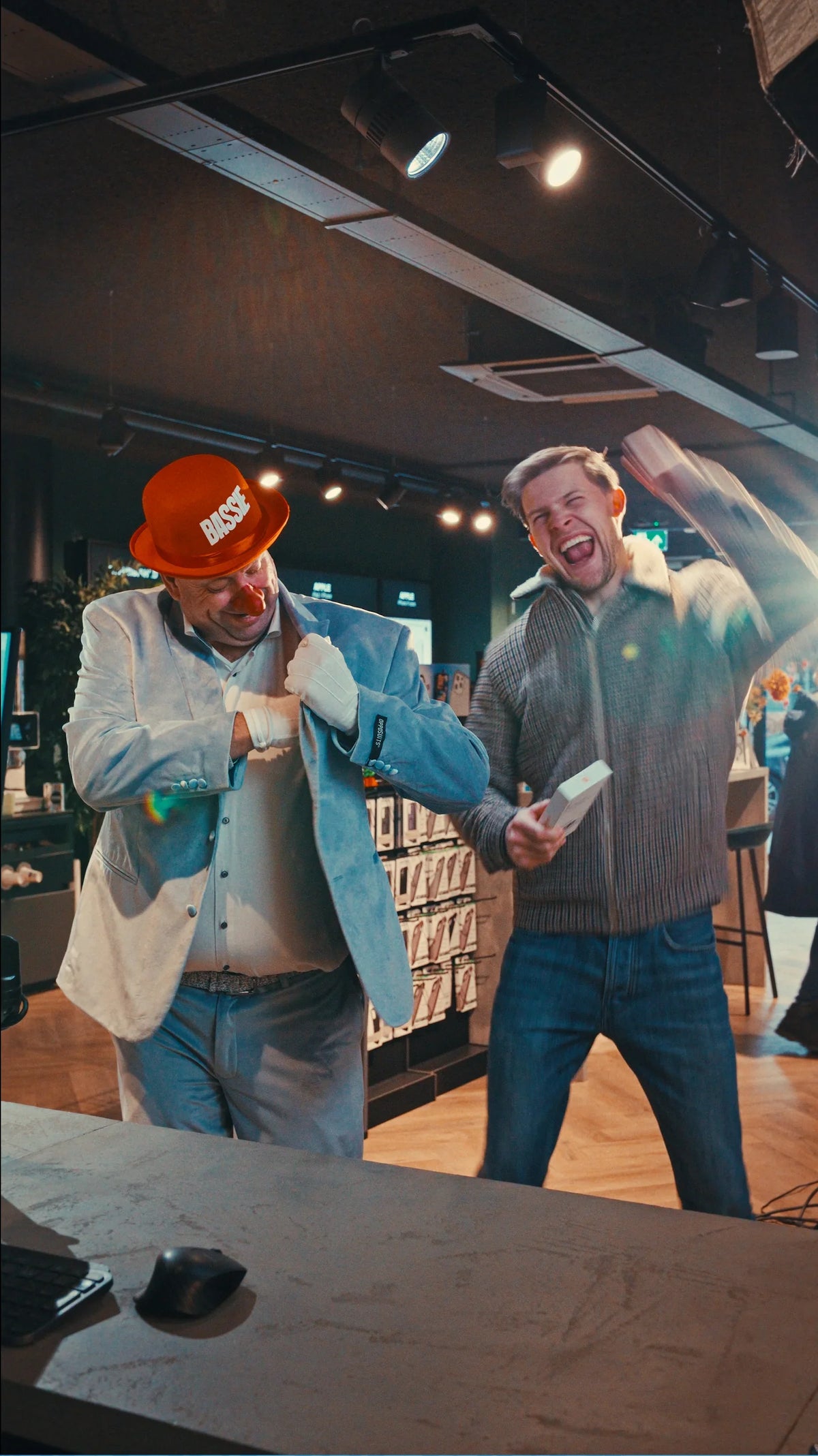 Twee blije mannen in elektronicawinkel, één met clownshoed en rode neus, ander met iPhone-doosje.